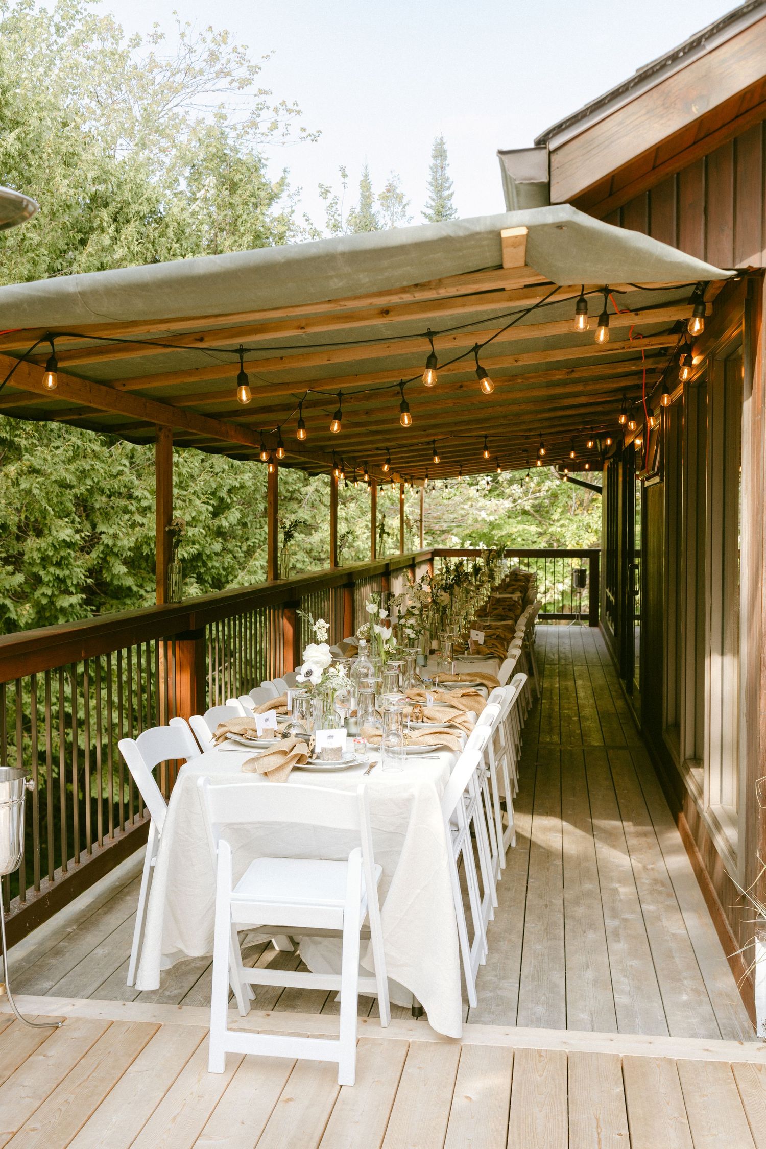 Elegant outdoor wedding reception table set on wooden deck with string lights and white chairs.