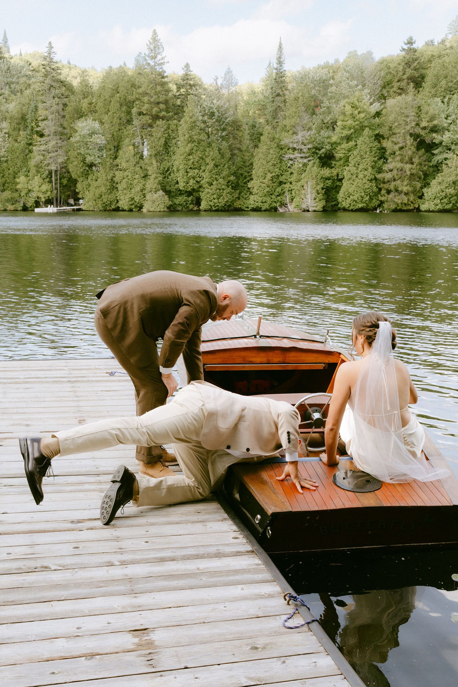 Groom and guest help bride board a classic wooden boat at a lakeside dock during a scenic wedding celebration.