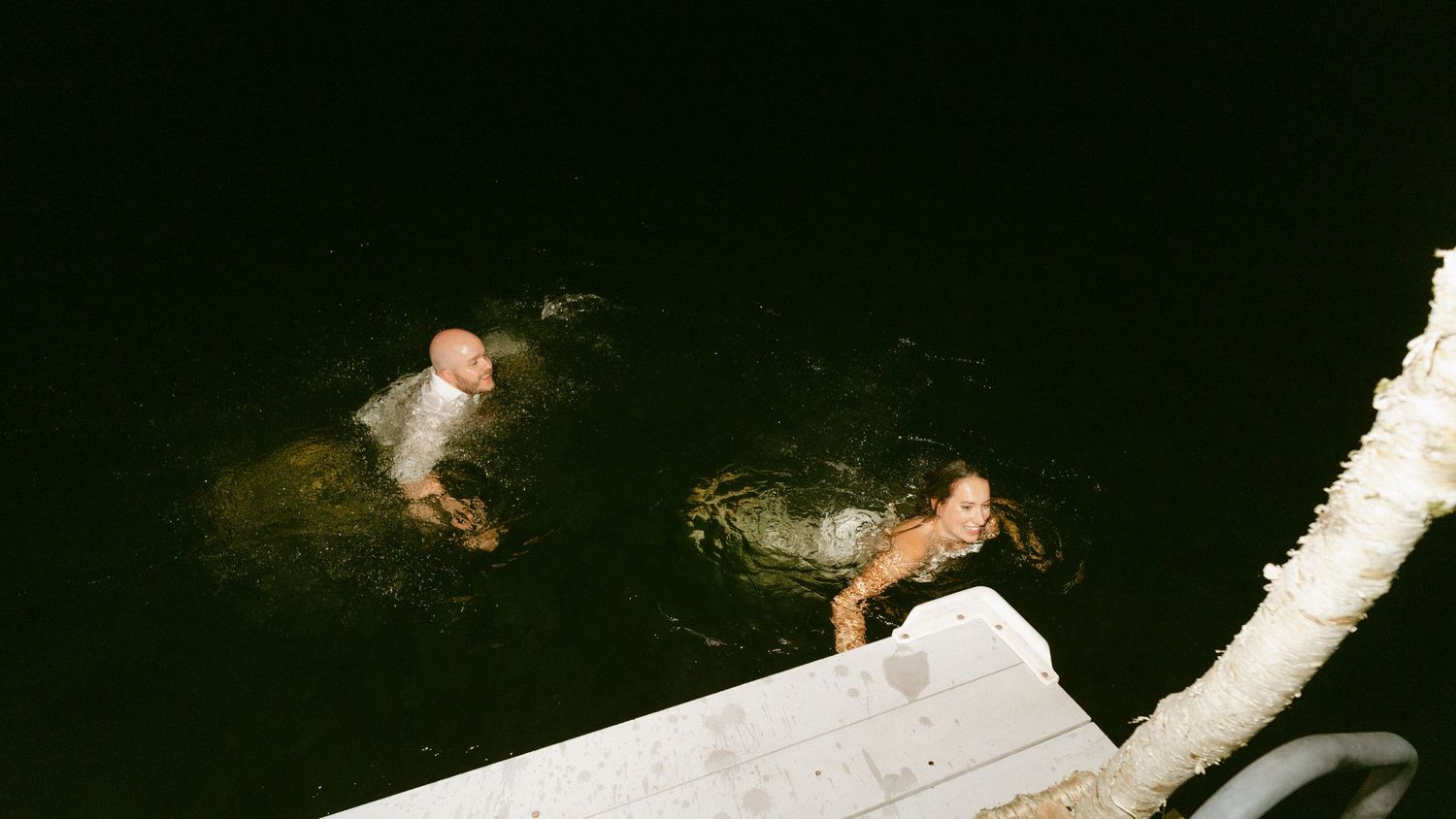 Bride and groom swimming in dark water at night after jumping off a dock in wedding attire.