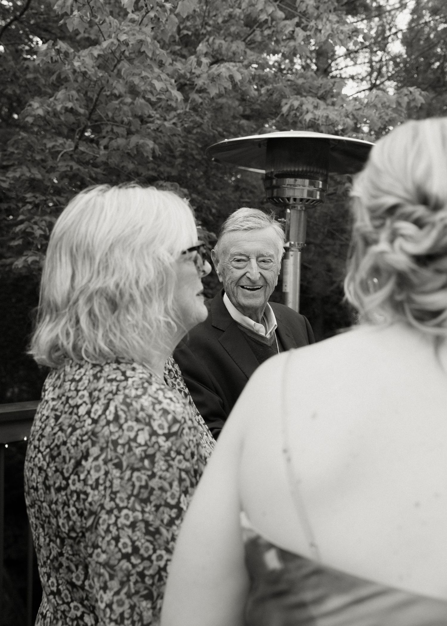Black and white photo of elderly man smiling warmly while conversing with guests at wedding.