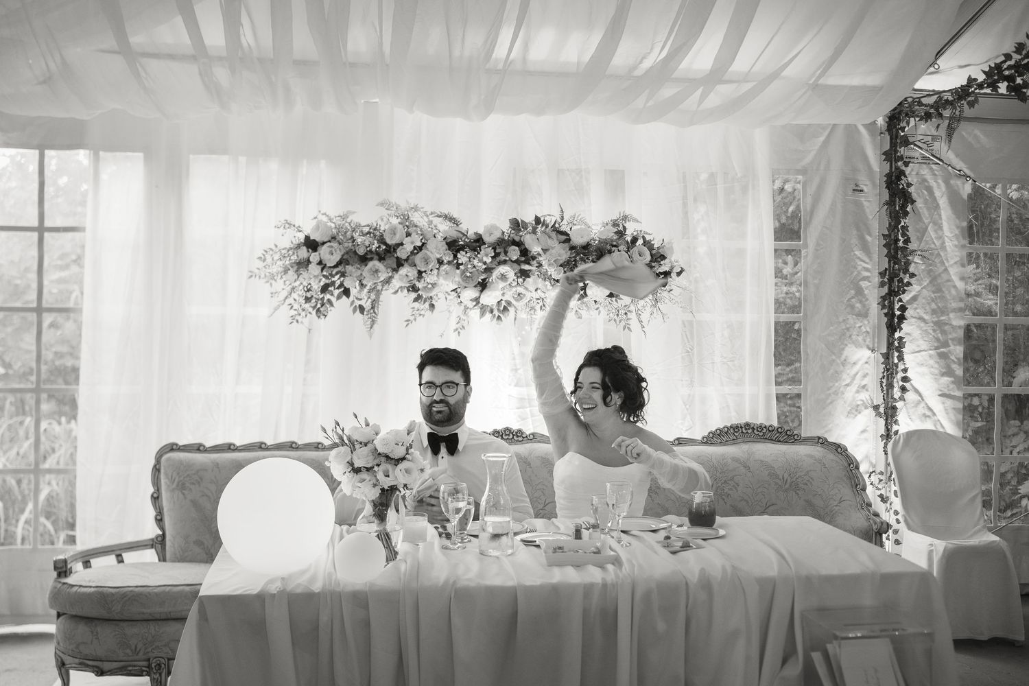 Black and white photo of bride raising arm joyfully at sweetheart table beneath floral arrangement.