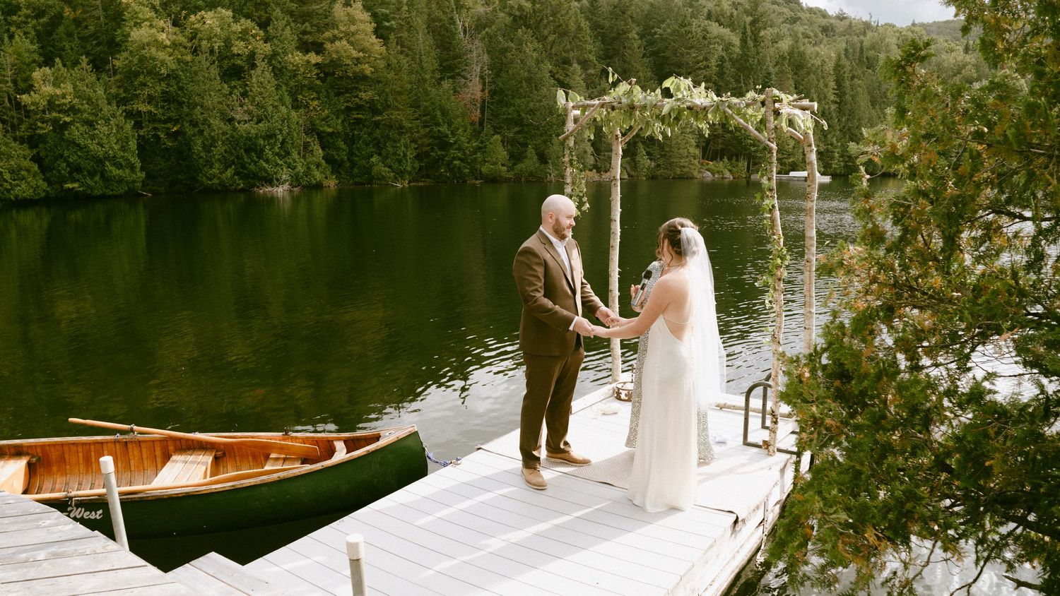 Bride and groom hold hands at a floral arch on a lake dock during their intimate ceremony.