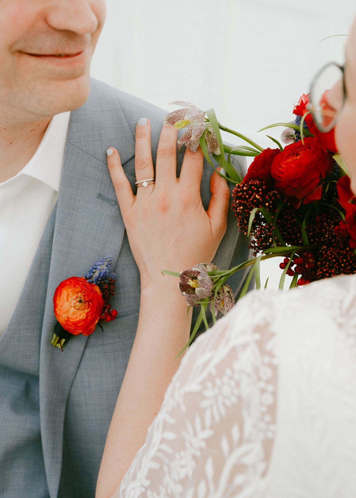 Close-up of bride resting hand on grooms gray suit lapel with red ranunculus boutonniere and bouquet.
