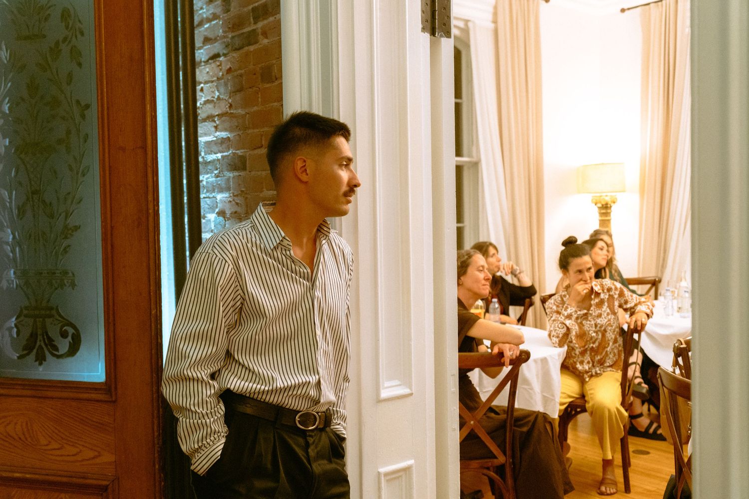 Man leaning against doorway observing wedding reception guests dining in background.