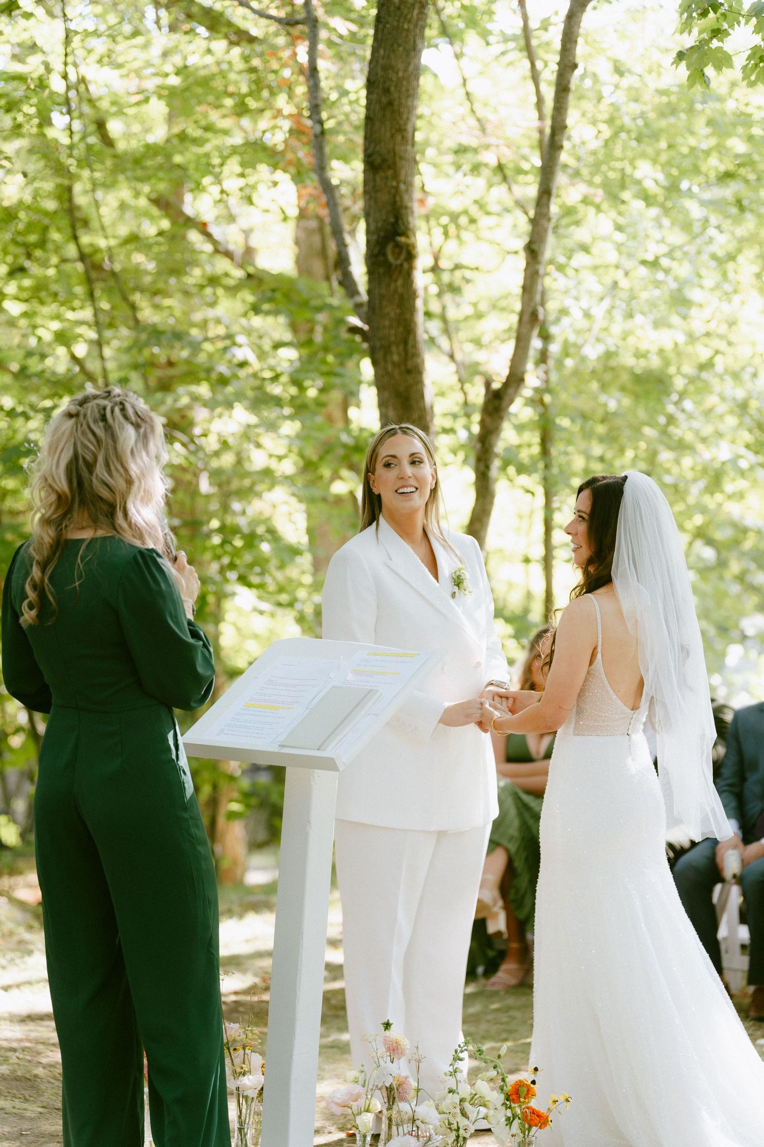 Two brides holding hands during an outdoor woodland wedding ceremony with an officiant.