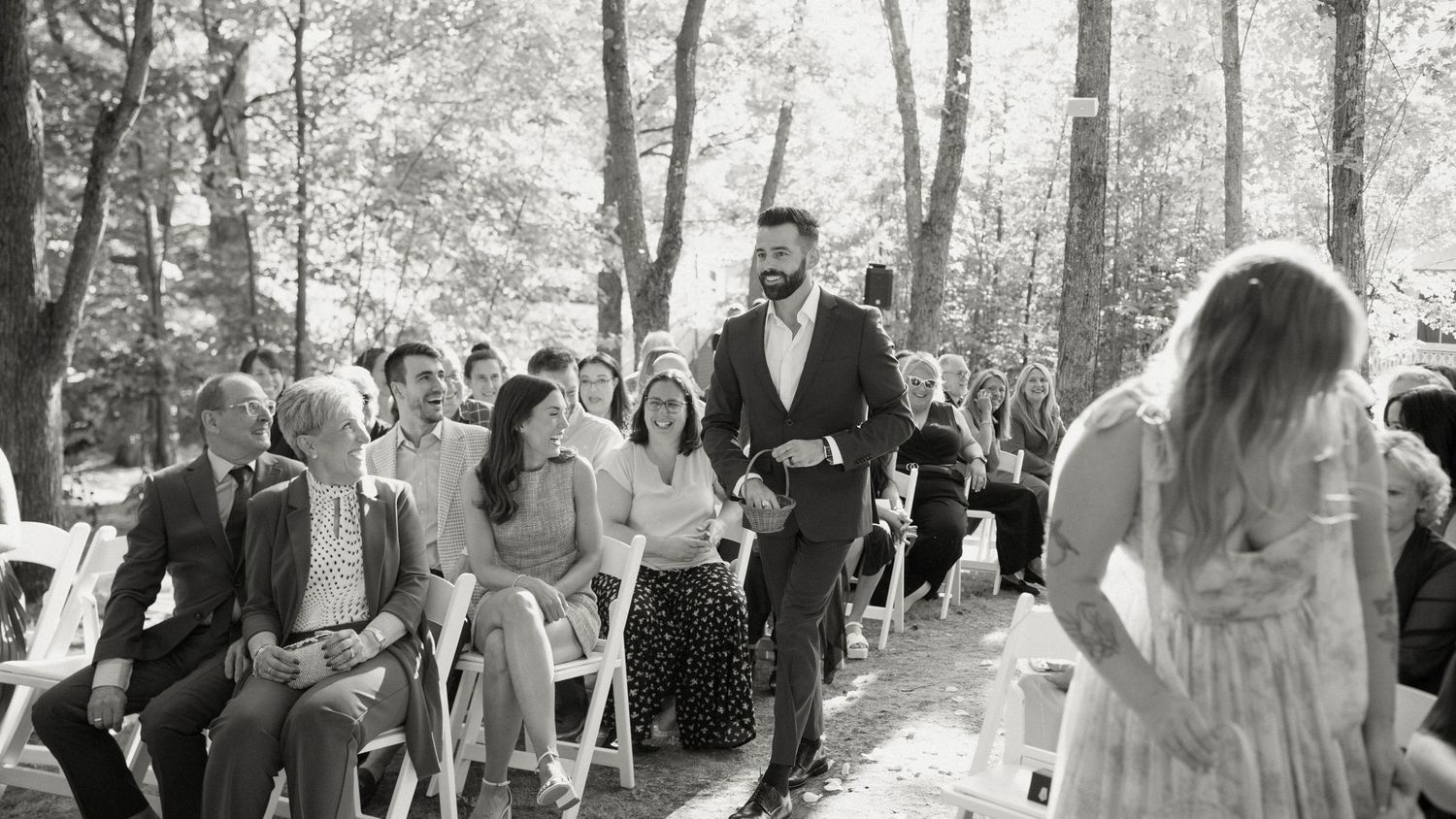 Bearded groomsman walks down tree-lined aisle as guests watch outdoor wedding ceremony.