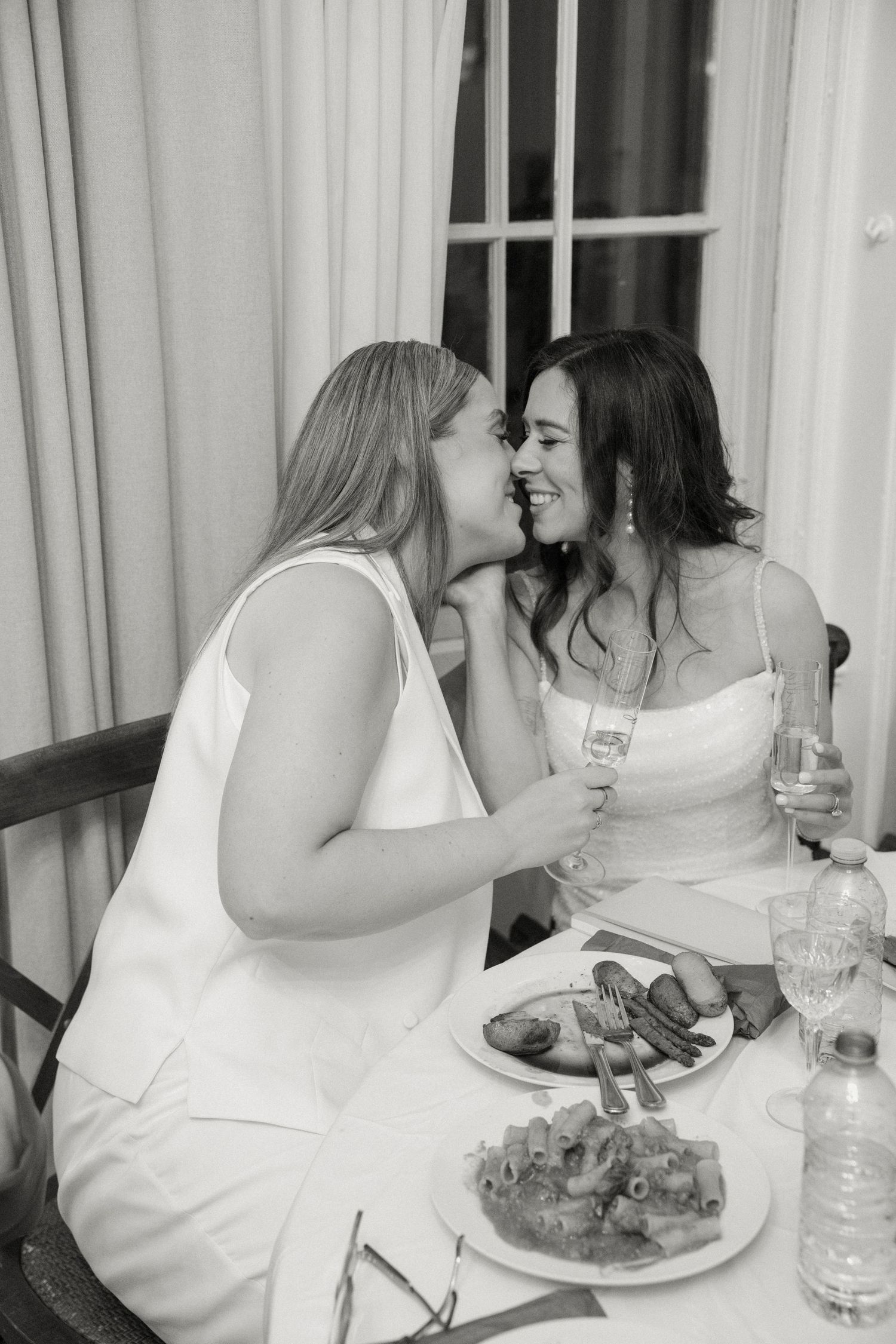 Two women in white dresses sharing a joyful moment at a wedding dinner table.