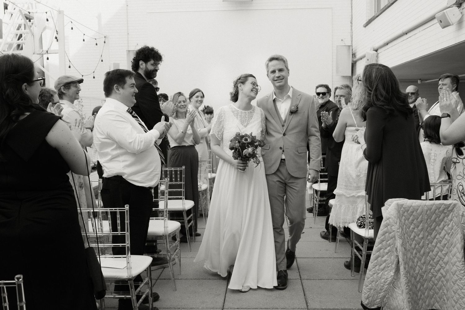 Bride and groom walk down the aisle in black and white photo, guests applauding at outdoor wedding.