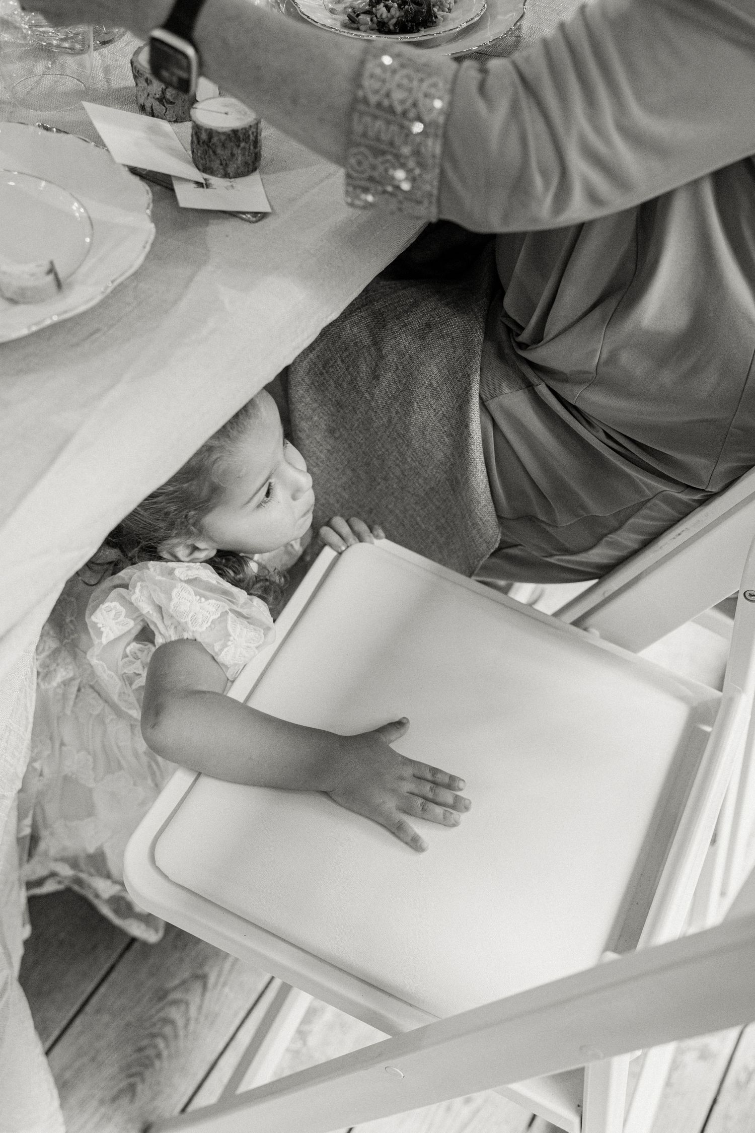Black and white photo of a young child playing under a wedding reception table in a white dress.