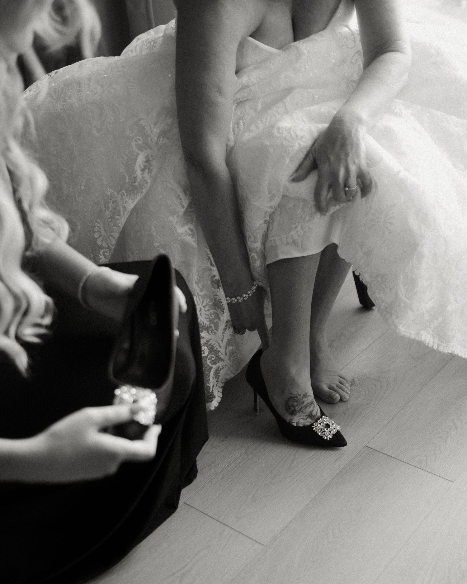 Bridesmaid helping bride put on decorative heels in black and white bridal preparation photo.