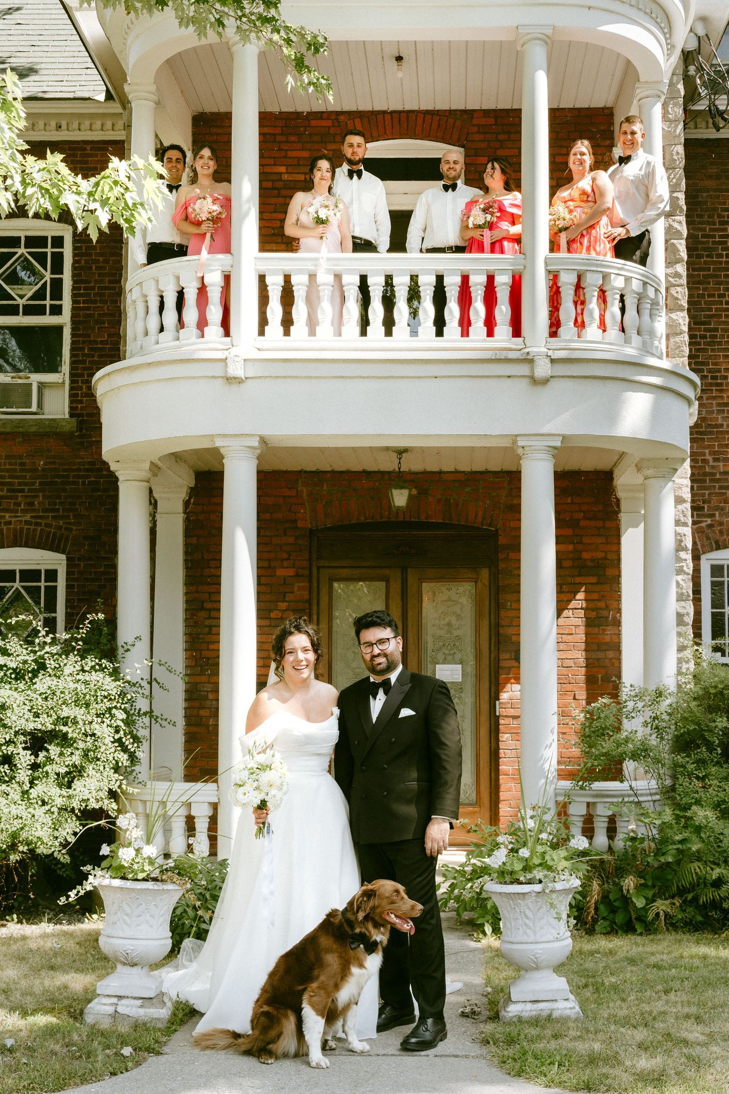 Wedding party poses on balcony as bride and groom stand with their dog below.
