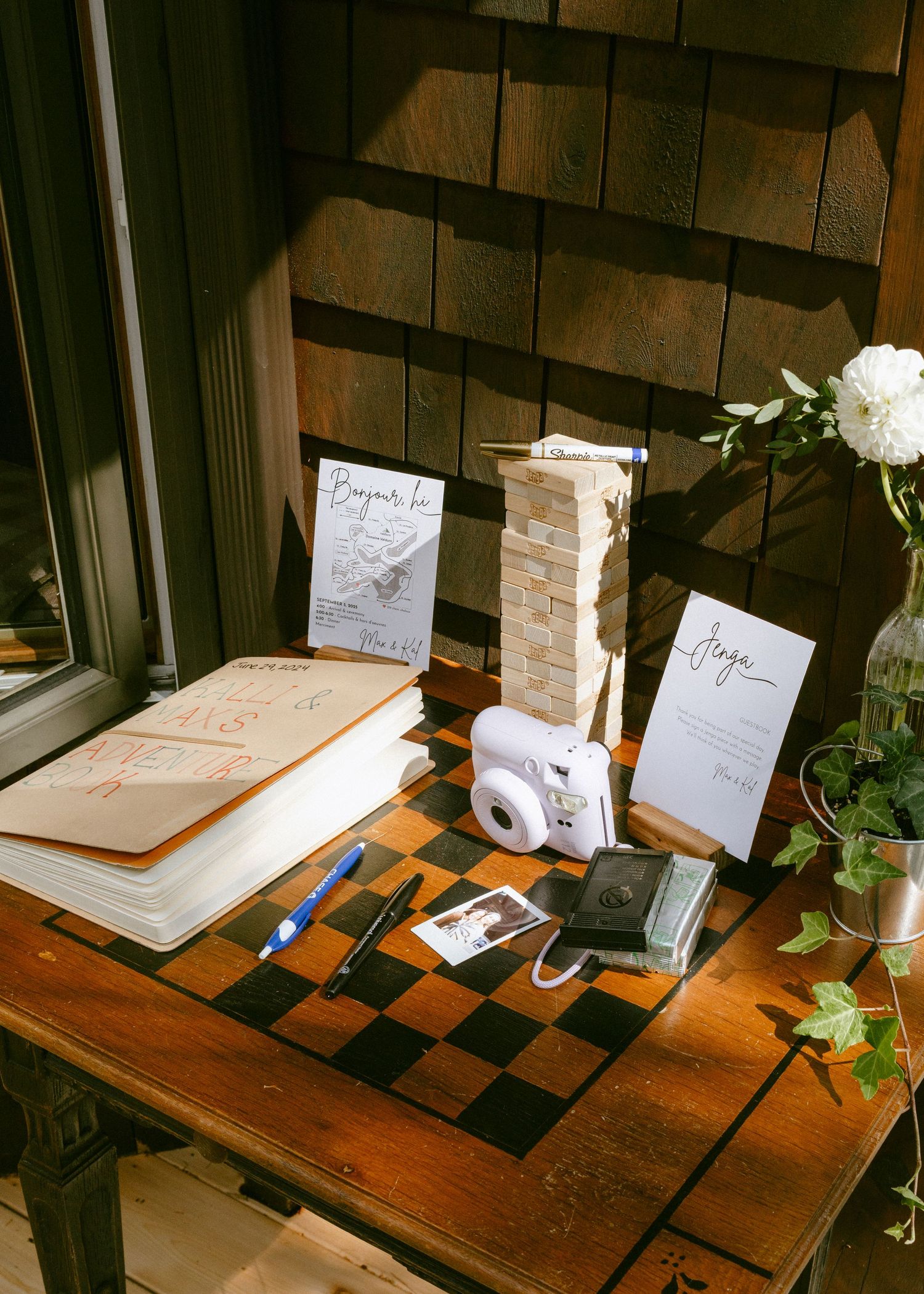 Rustic wedding guest book table with Instax camera, Jenga tower, guestbook, and white floral arrangement on checkered surface.