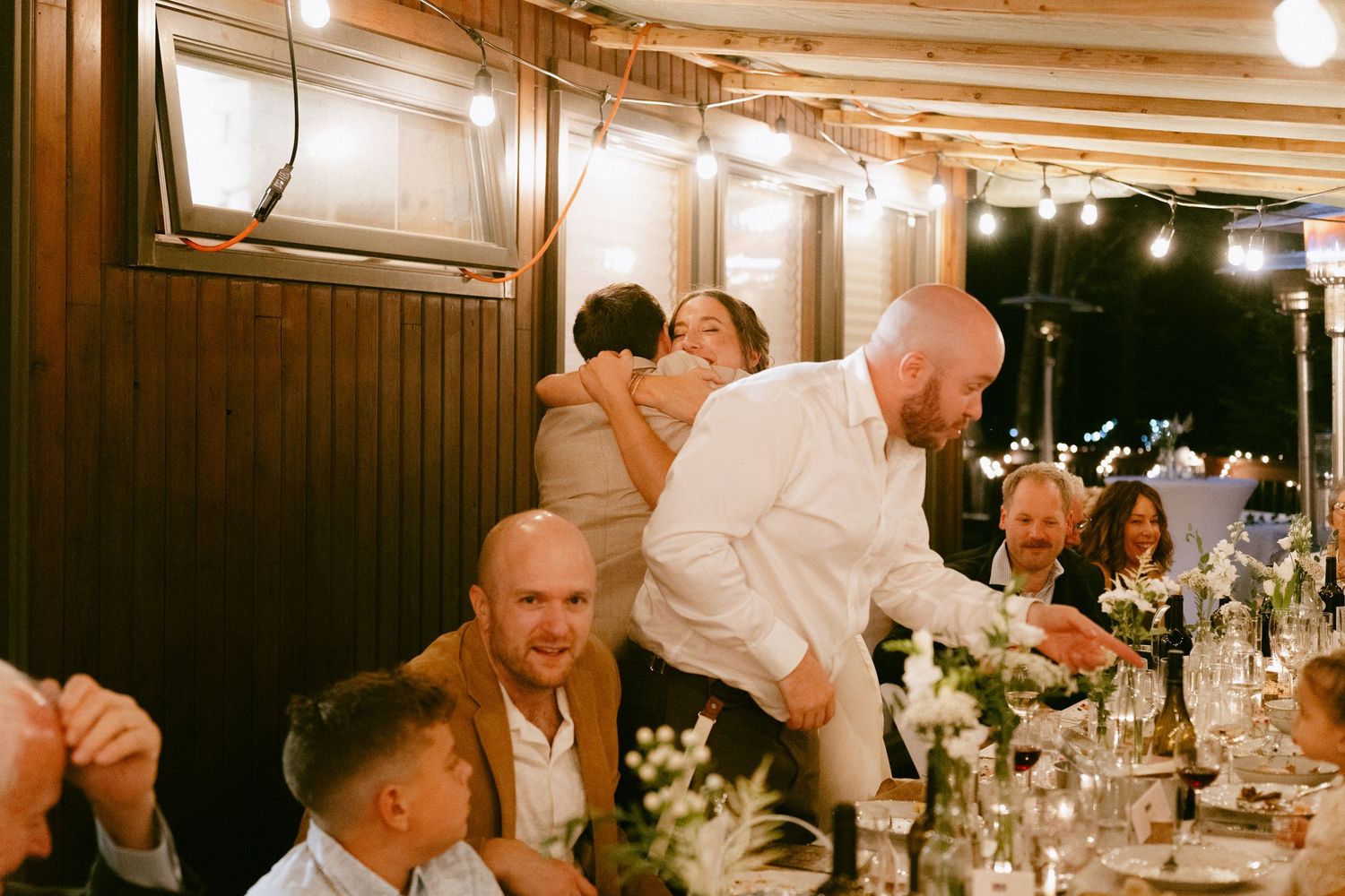 Bald man in white shirt embracing guests at a warmly lit wedding reception dinner.