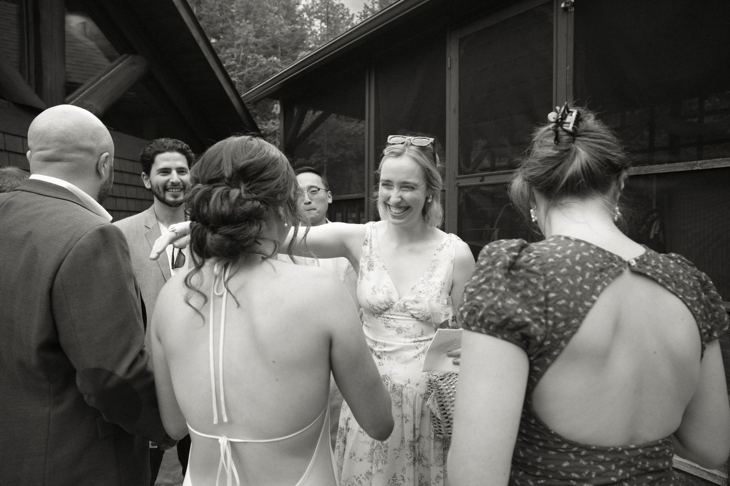 Black and white photo of wedding guests laughing and embracing outdoors near a wooden building.