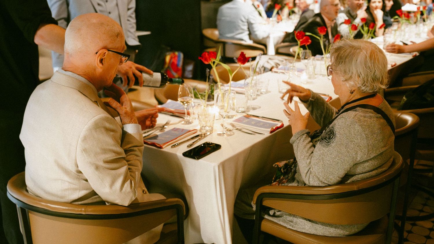 Two wedding guests engaged in conversation at a decorated reception table with red floral centerpieces.