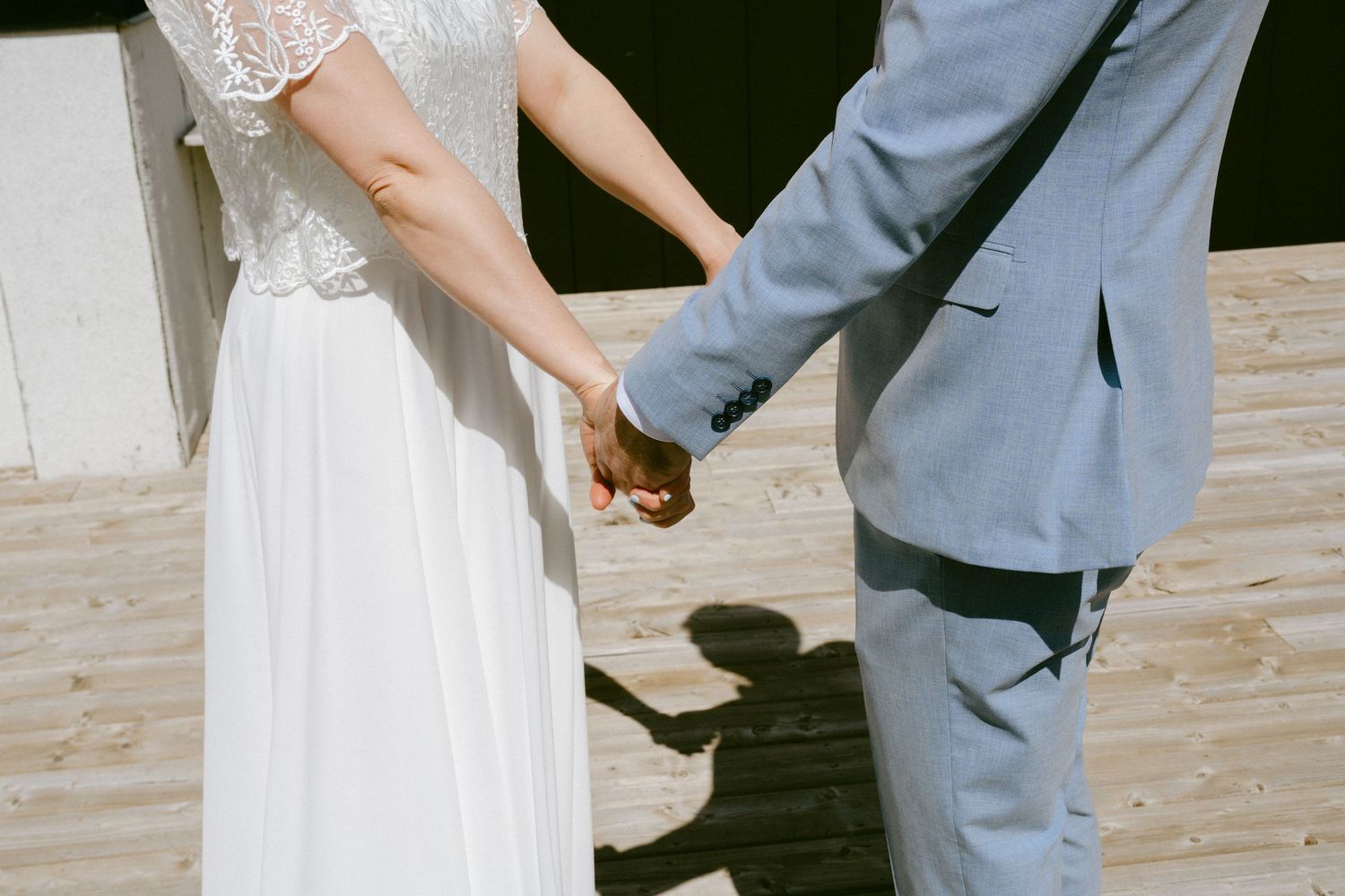 Close-up of bride and groom holding hands casting shadows on a sunny wooden deck.
