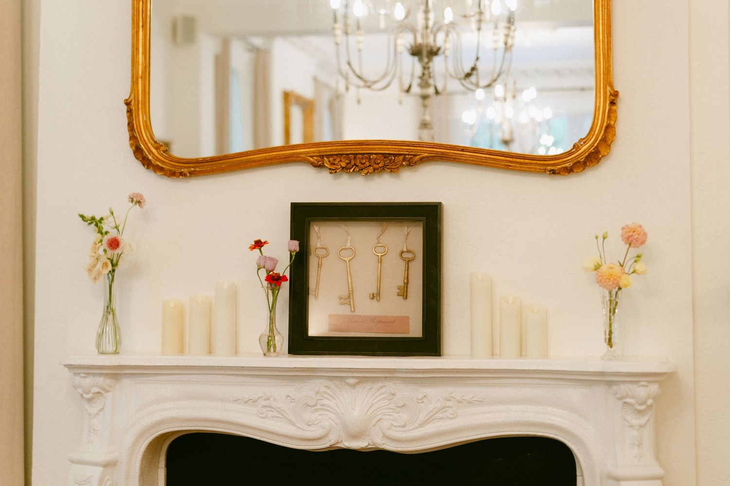Elegant white fireplace mantel with gold mirror, candles, and delicate floral arrangements in glass vases.