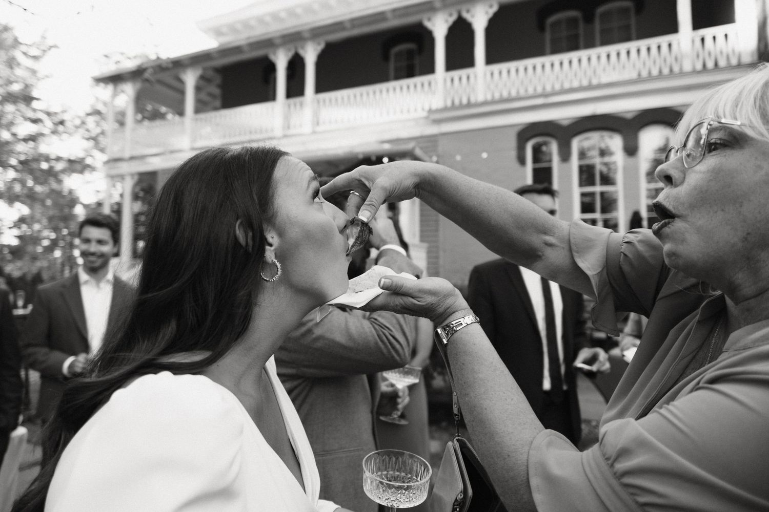 Black and white photo of a woman being fed a bite of food at an outdoor wedding reception.