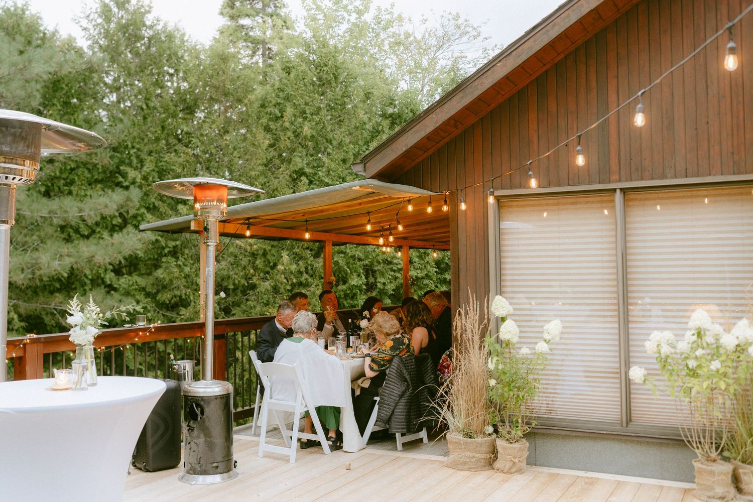 Guests dining on a wooden deck with string lights and patio heaters surrounded by trees.