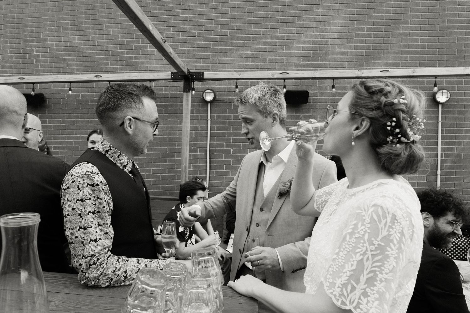 Bride with floral hair accessory converses with guests in patterned vest at wedding bar.