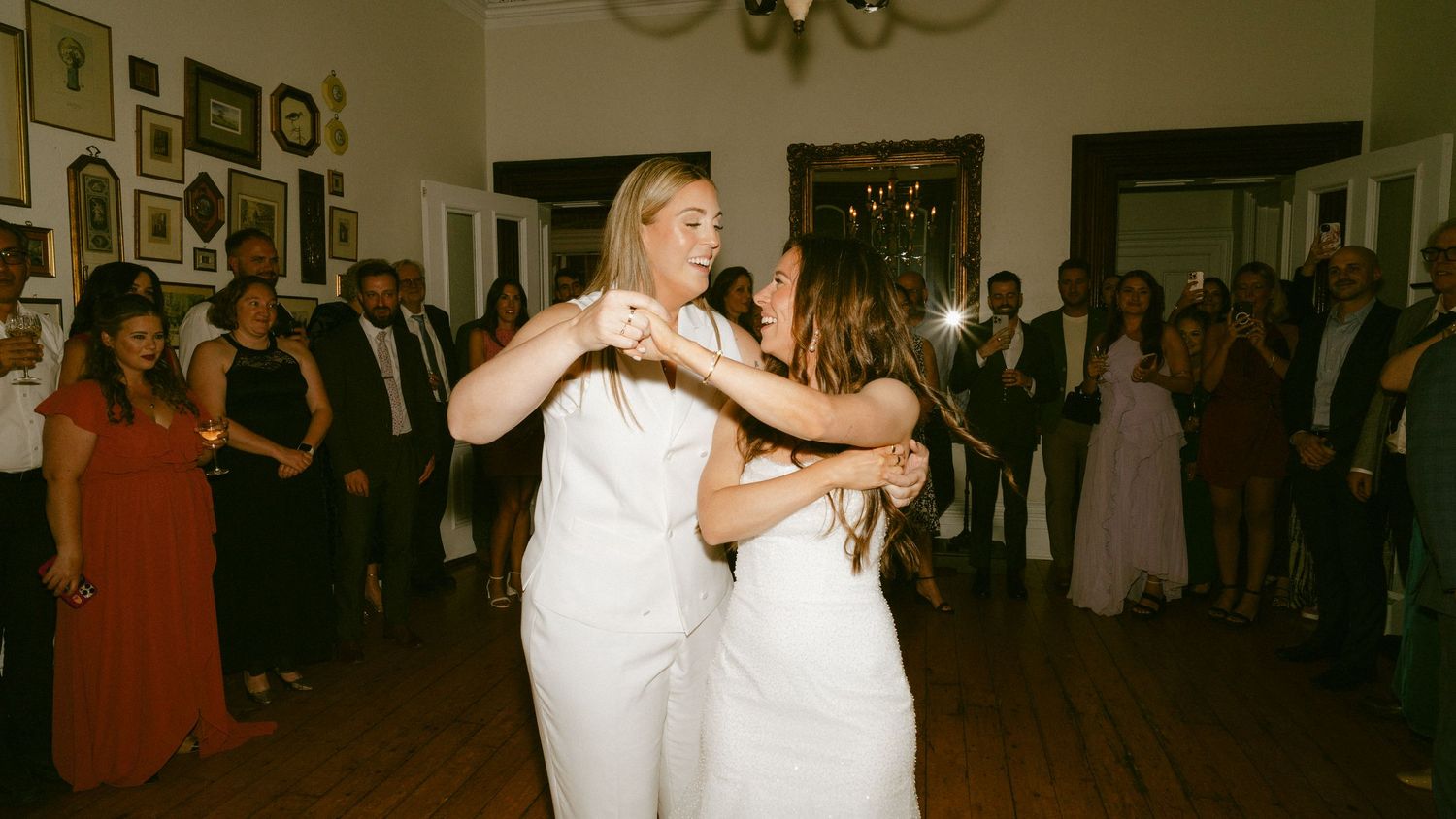 Two brides in white share a joyful first dance surrounded by wedding guests in elegant venue.