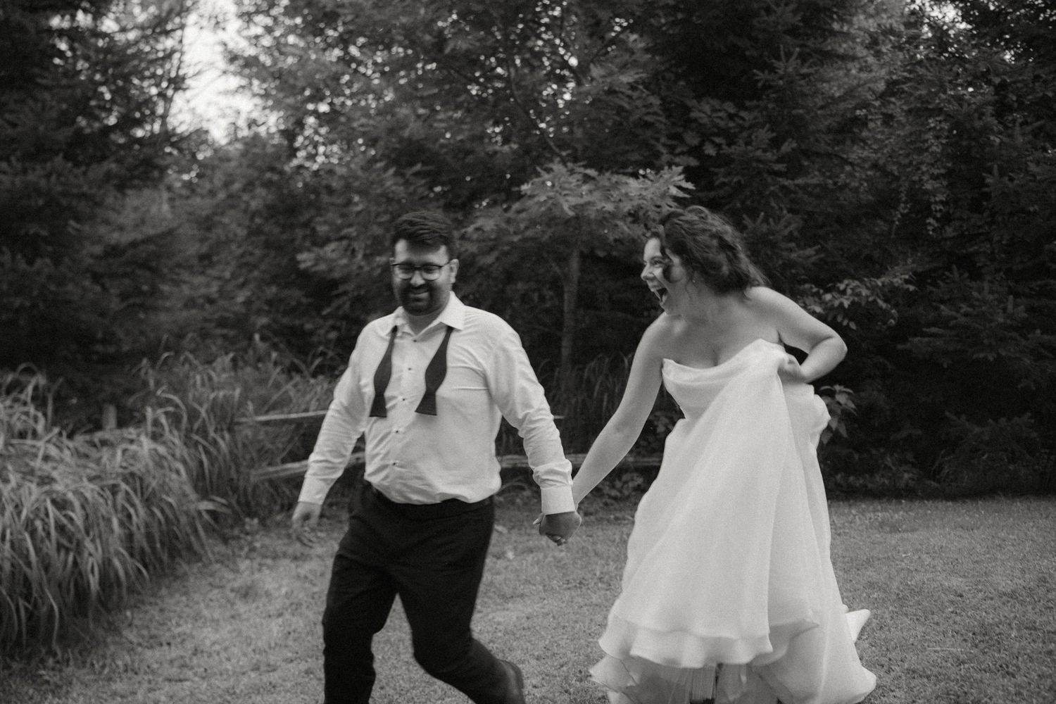 Black and white photo of laughing bride and groom running playfully across green lawn together.