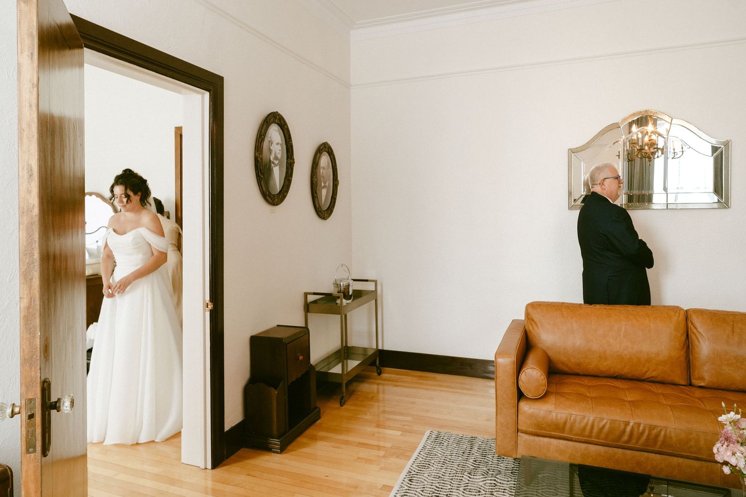 Bride in white gown stands in doorway as suited man waits with back turned for first look reveal.
