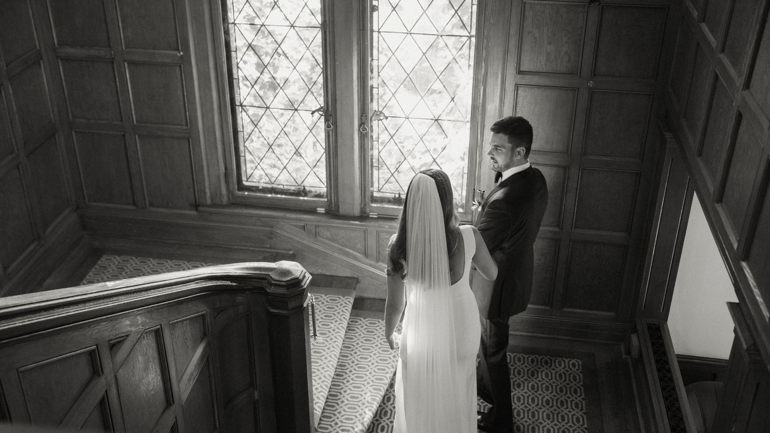 Black and white photo of groom adjusting brides veil near a stained glass window on wedding day.