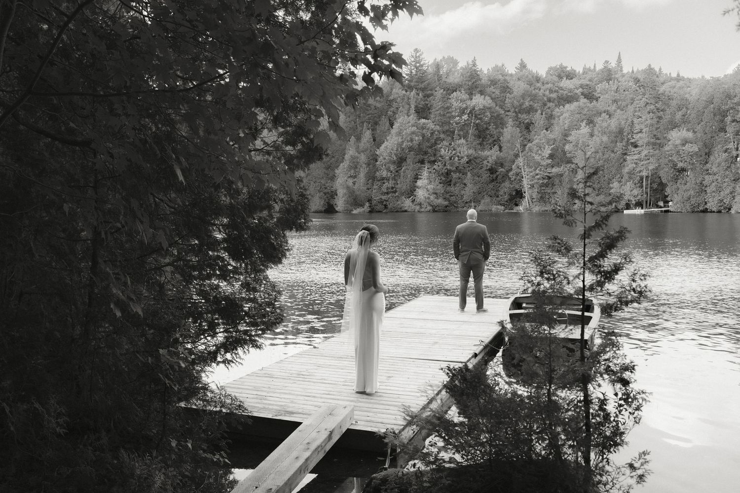 Bride and groom stand apart on a wooden dock surrounded by a serene forest lake.