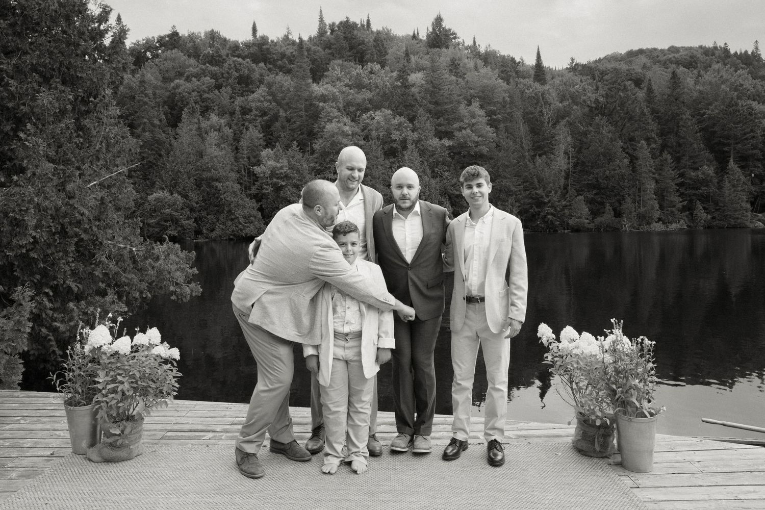 Group of five men posing together on a lakeside dock surrounded by lush forest in a black and white wedding photo.