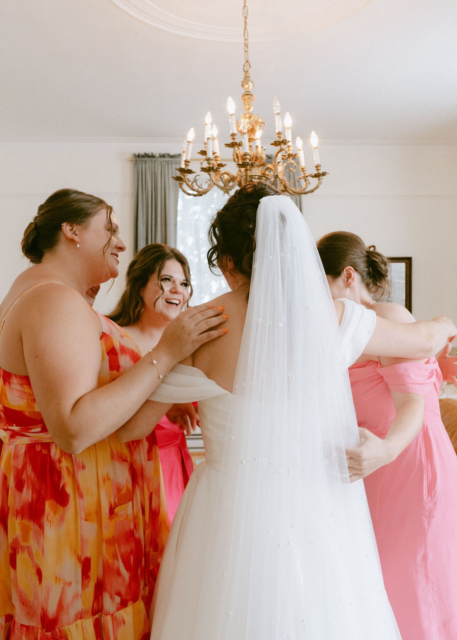 Bridesmaids in coral dresses surround smiling bride in white wedding gown beneath golden chandelier.