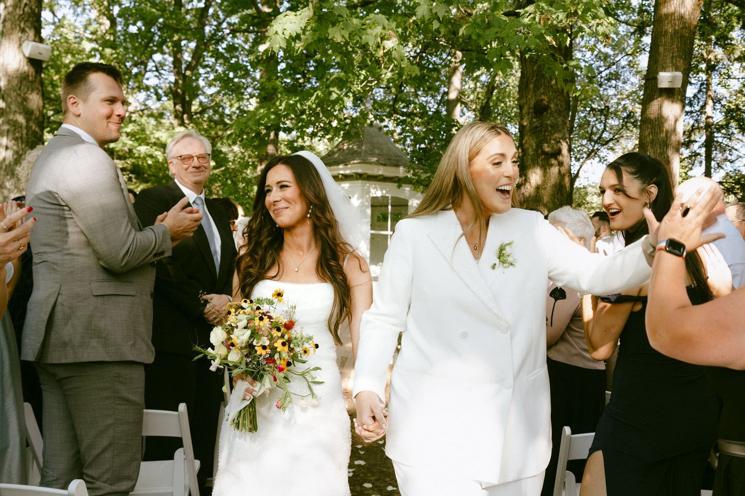 Two brides walk hand-in-hand down the aisle as guests celebrate their outdoor wedding ceremony.