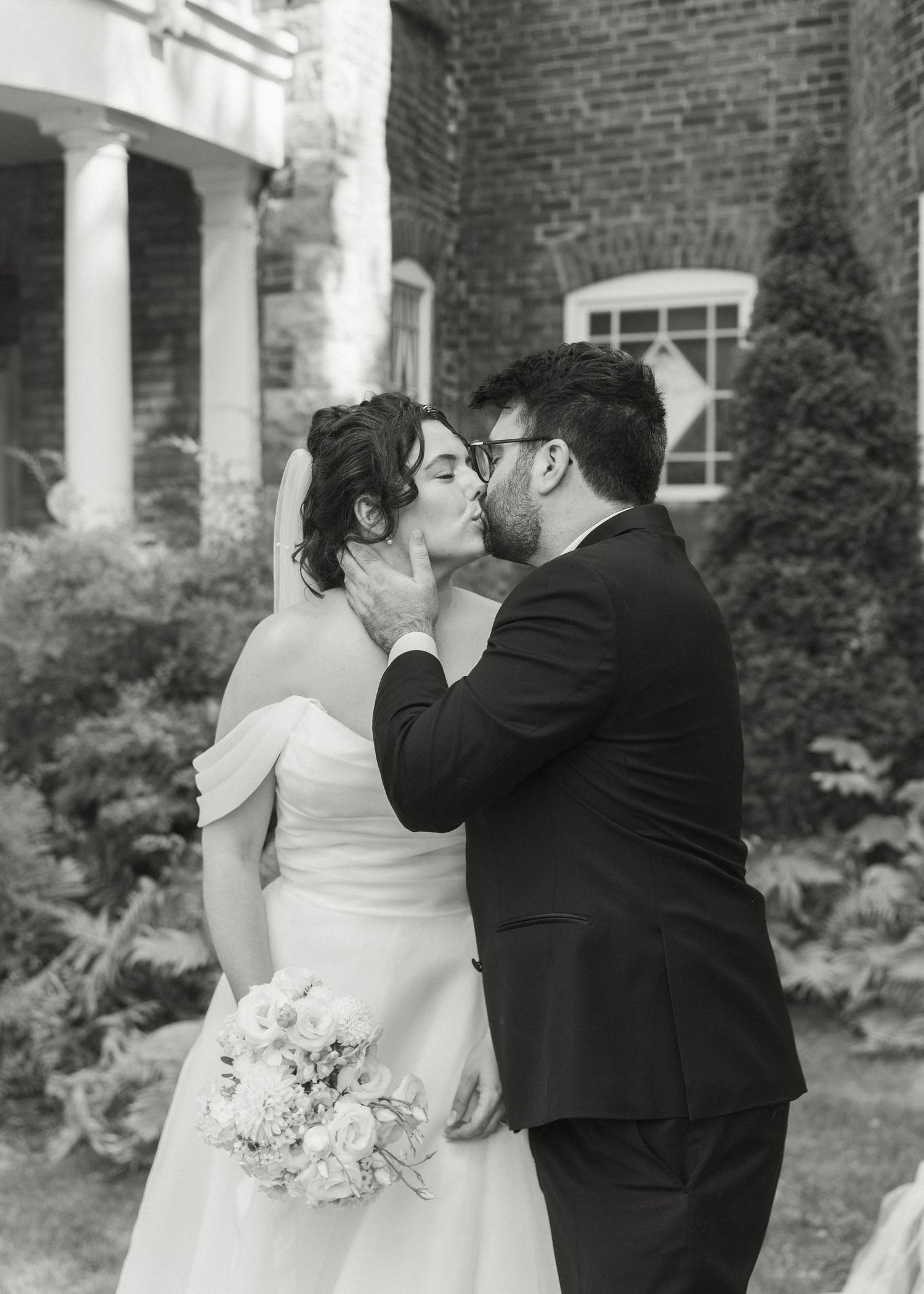Bride and groom share a romantic kiss in black and white photo outside brick venue.