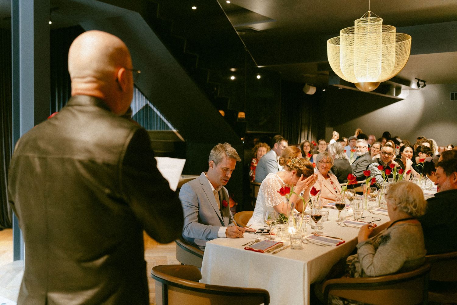 Bald man in leather jacket addressing wedding guests seated at long tables in an elegant restaurant venue.