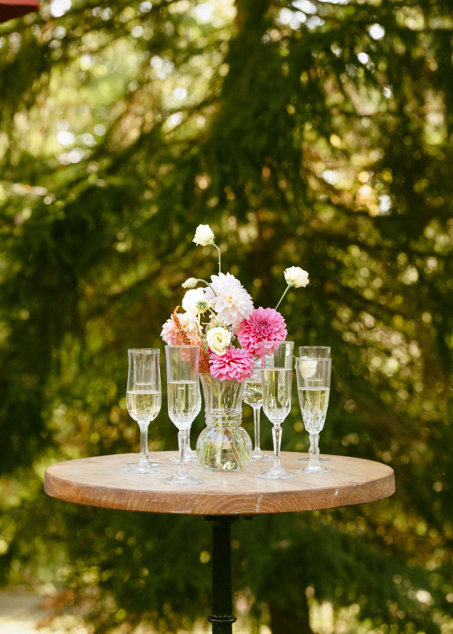 Round wooden cocktail table with champagne flutes and pink dahlia floral centerpiece outdoors.