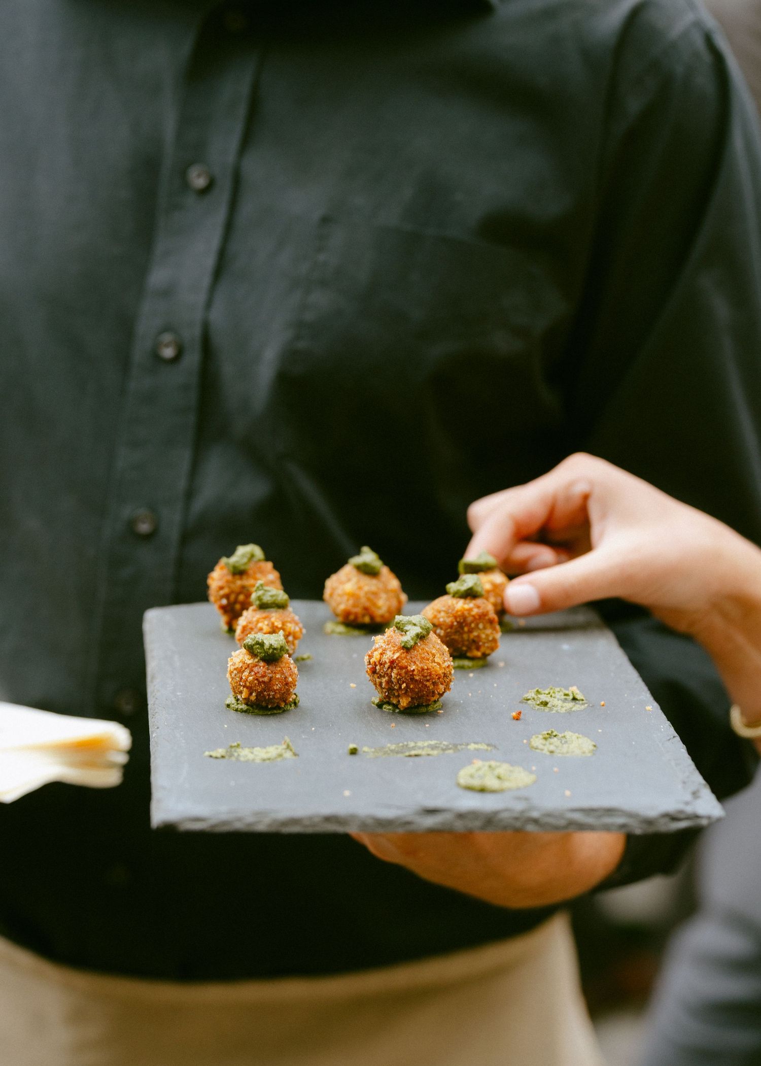 Catering server presenting breaded appetizer bites garnished with herbs on a slate serving board.