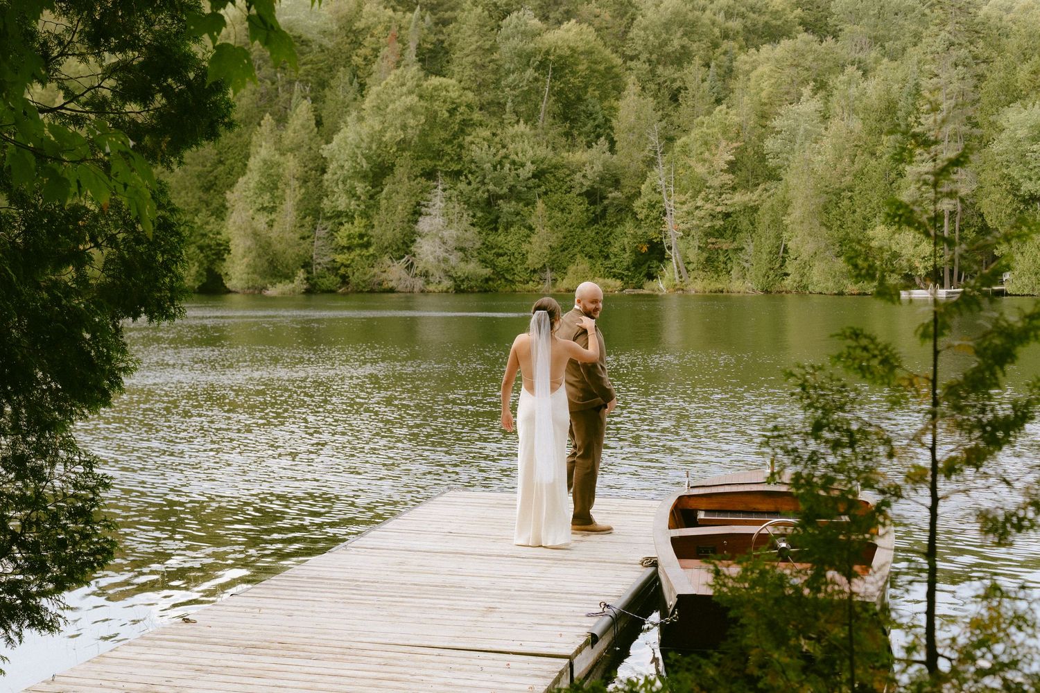 Bride and groom embracing on a wooden dock beside a calm lake surrounded by green forest trees.