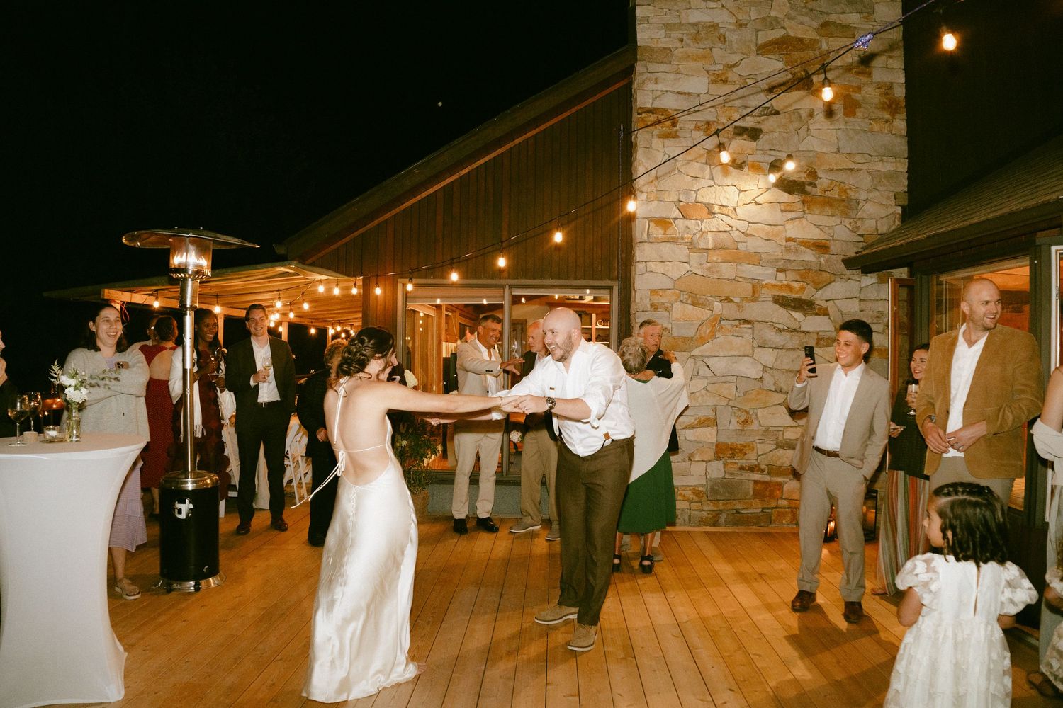 Bride in white gown dancing with older man on outdoor deck at night wedding reception.