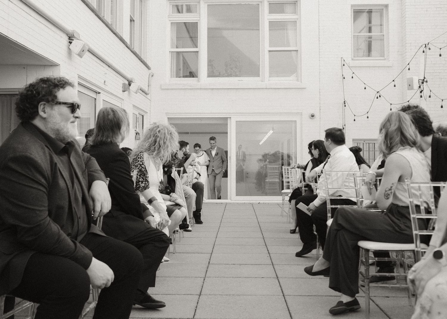 Black and white photo of wedding guests seated in an outdoor courtyard watching a ceremony at the altar.