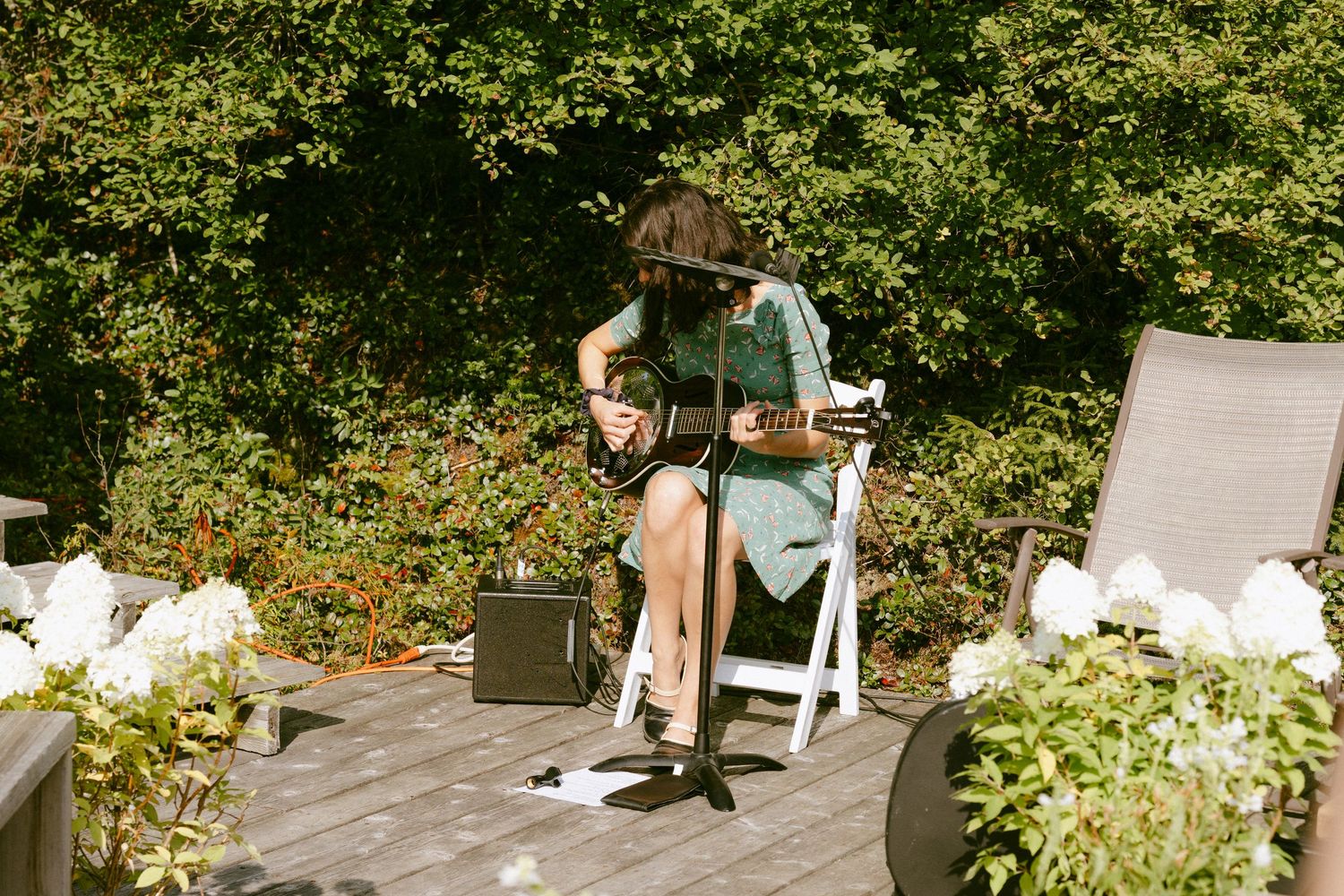 Musician playing guitar on a sunny outdoor deck surrounded by lush greenery and white flowers.