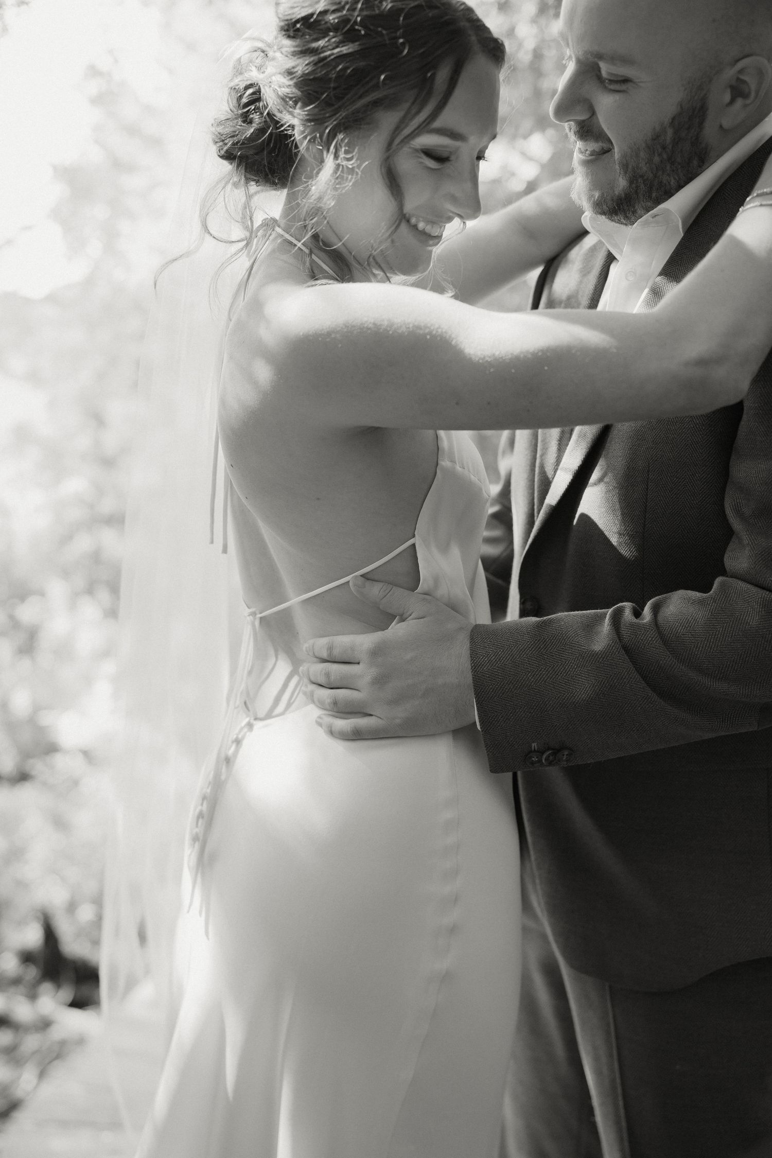 Black and white photo of laughing bride in open-back gown embraced by bearded groom in dappled forest light.