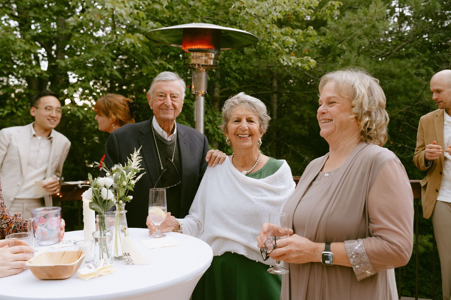 Smiling wedding guests gathered around cocktail table on outdoor deck surrounded by lush greenery.