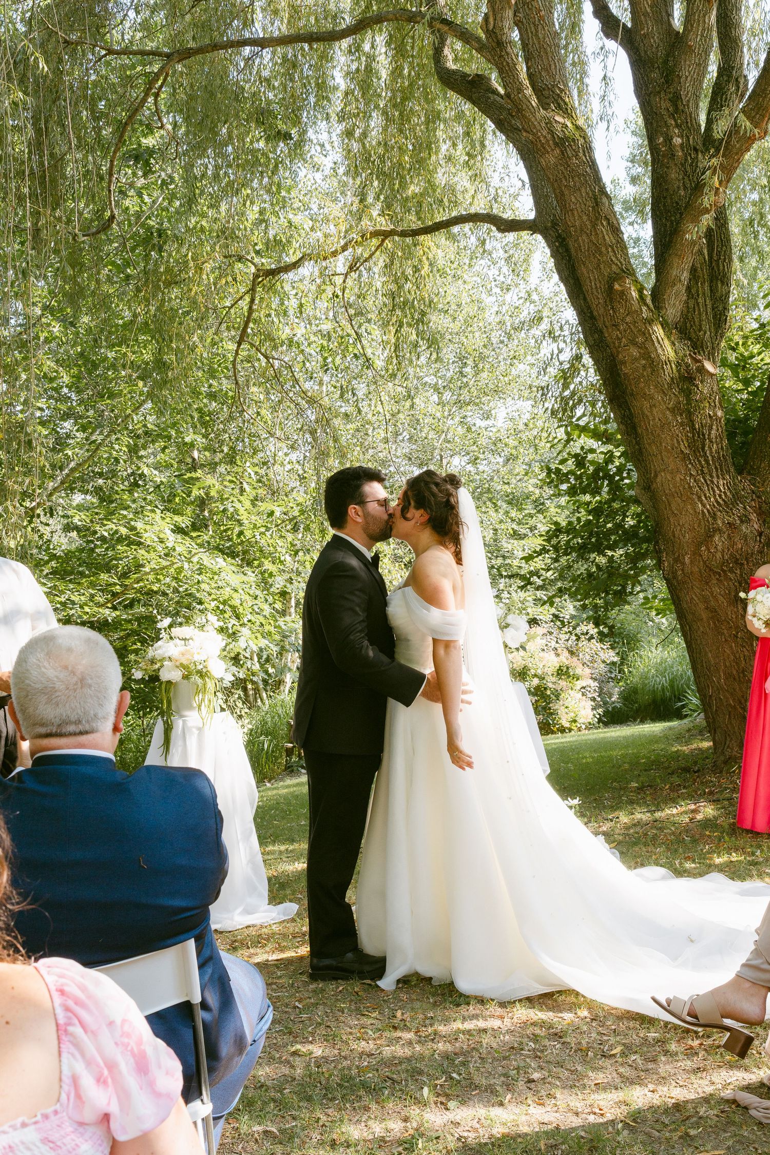 Bride and groom share first kiss under willow tree at outdoor wedding ceremony.