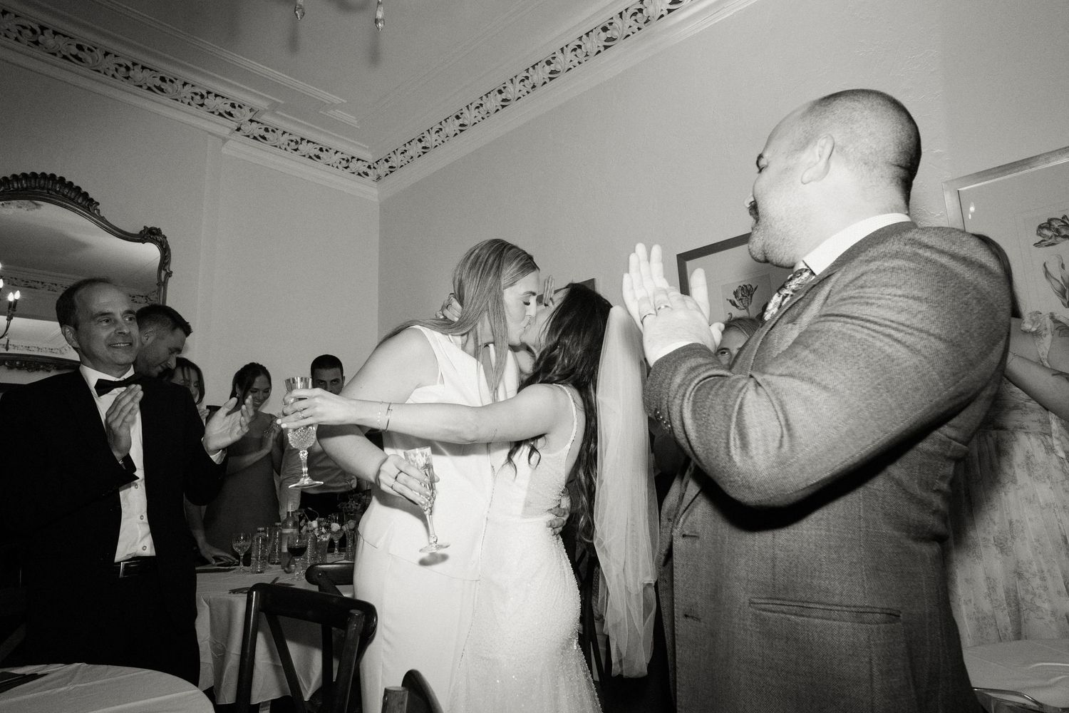 Two brides sharing a kiss while wedding guest photographs the moment in black and white.