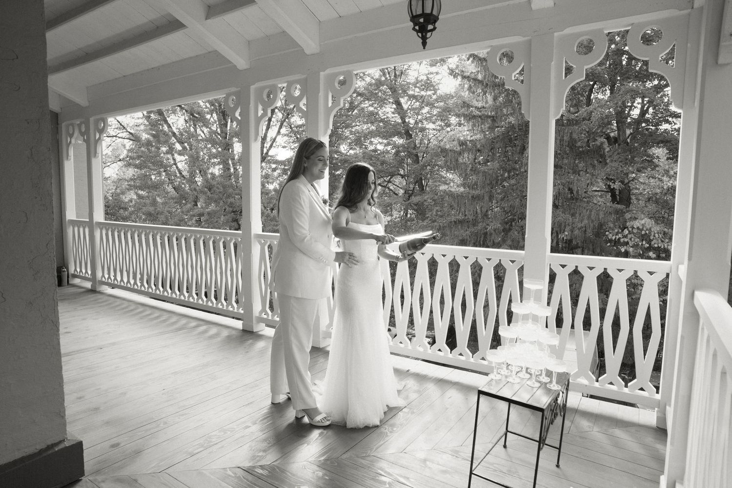 Two brides in white share vows on a Victorian porch in a black and white wedding photo.