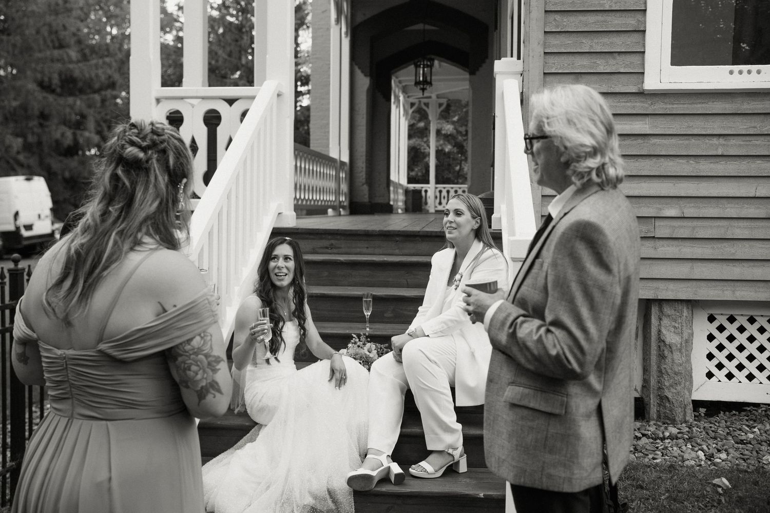 Bride in white gown and partner in white suit chat with guests on decorated porch steps at wedding.