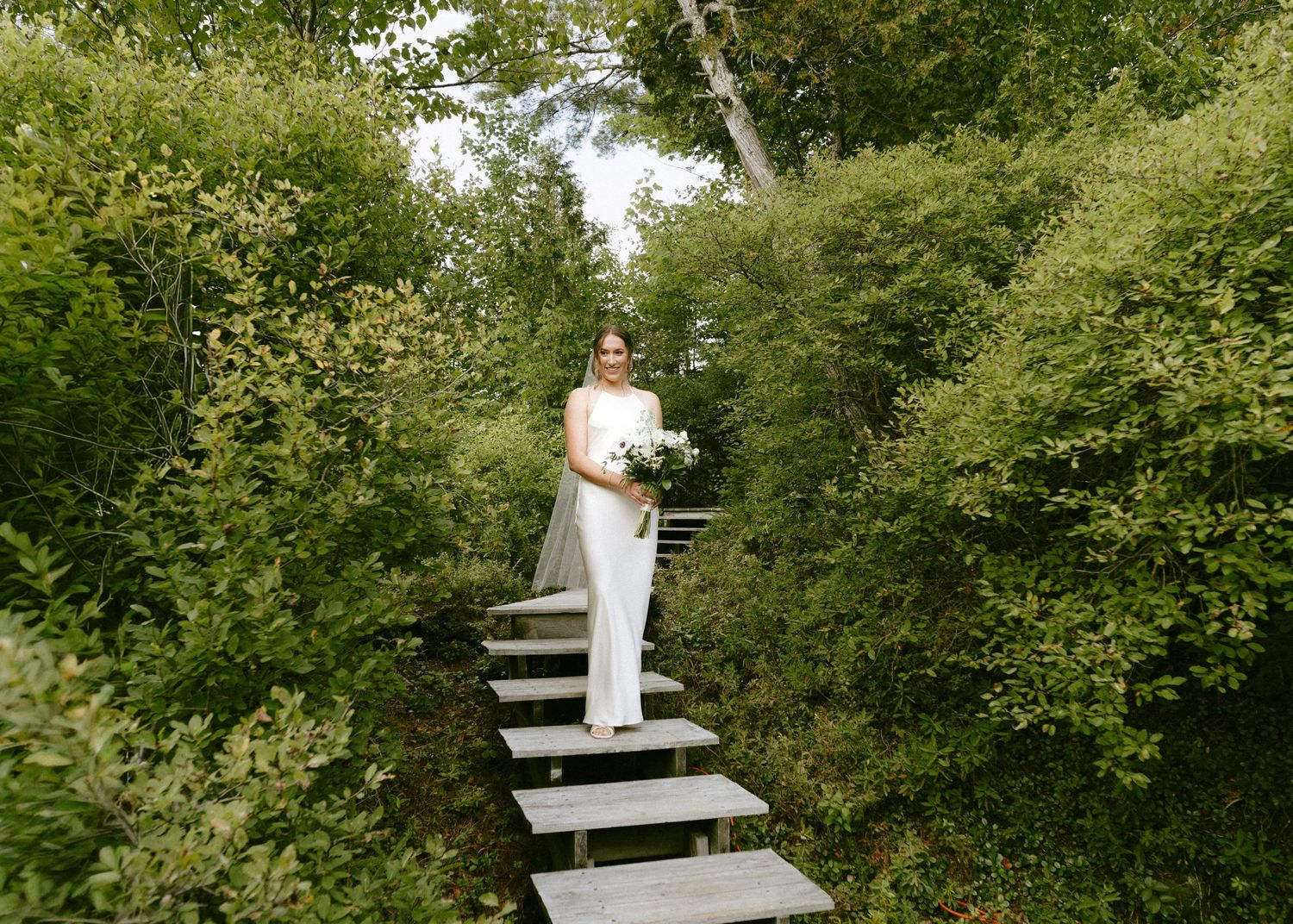 Elegant bride in white gown holding bouquet while standing on steps surrounded by greenery.