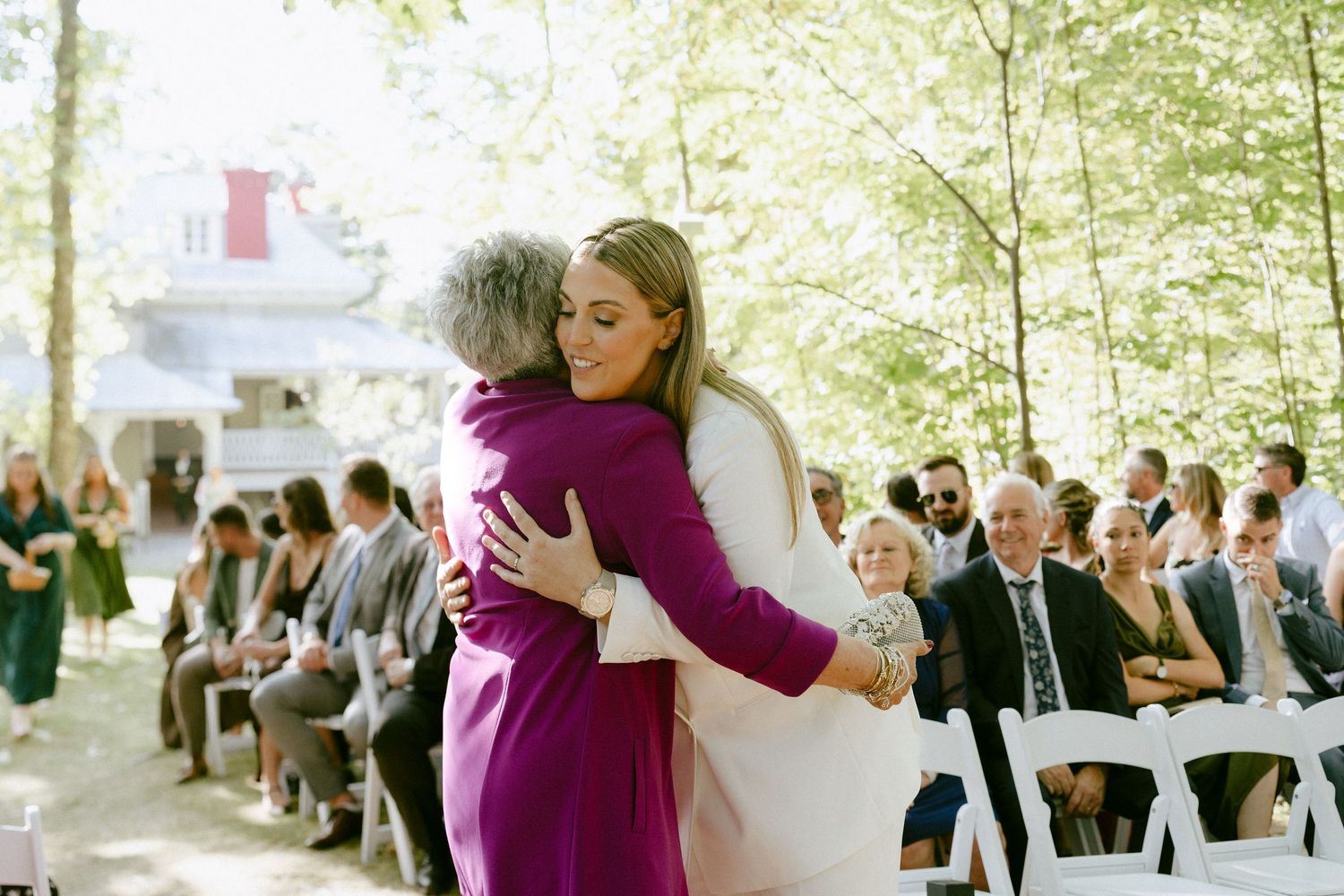 Bride in white embraces woman in purple suit during emotional outdoor wedding ceremony moment.