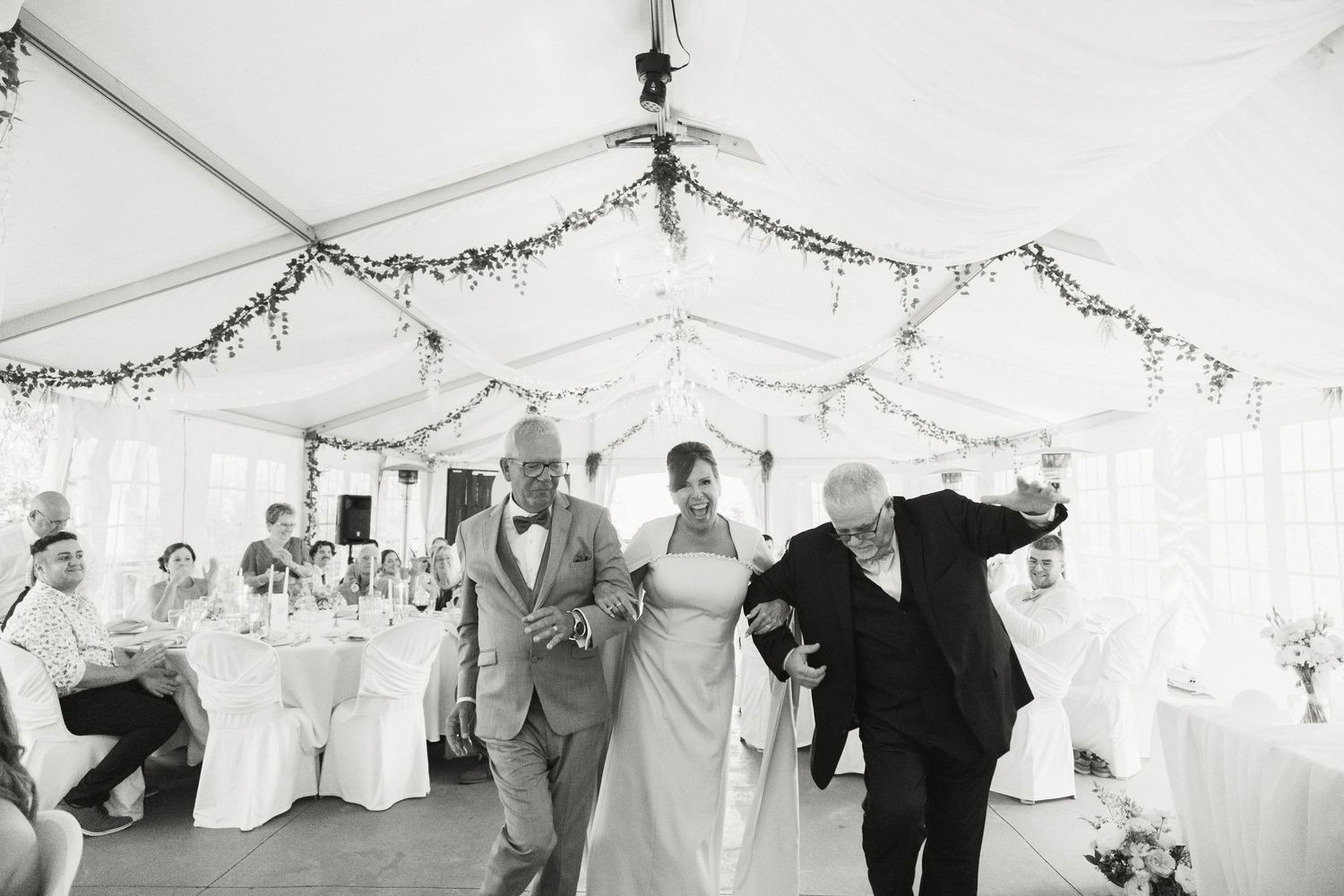 Black and white photo of bride and groom grand entrance into decorated wedding reception tent with guests.