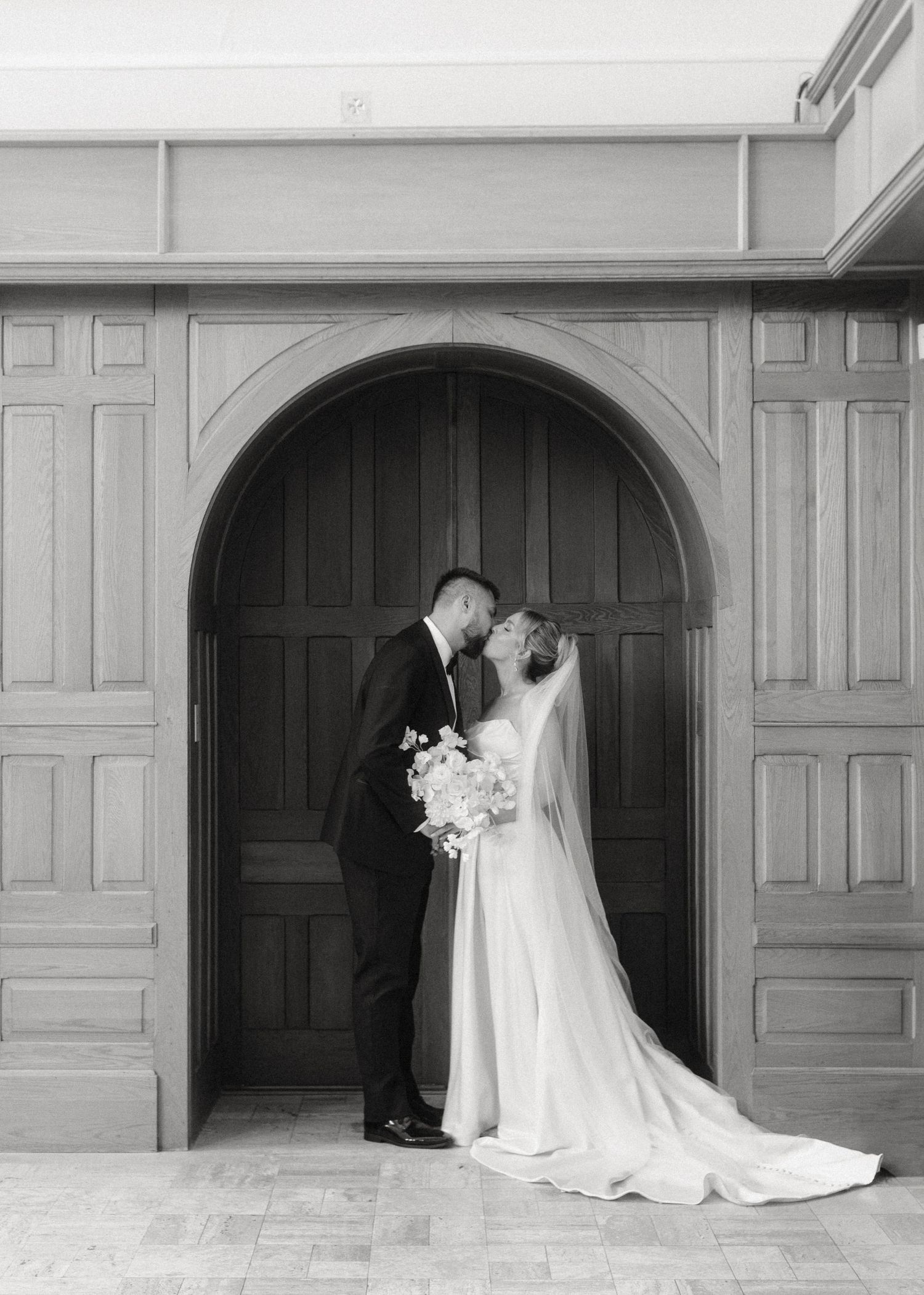 Bride and groom share a romantic kiss framed by a grand wooden arched doorway.
